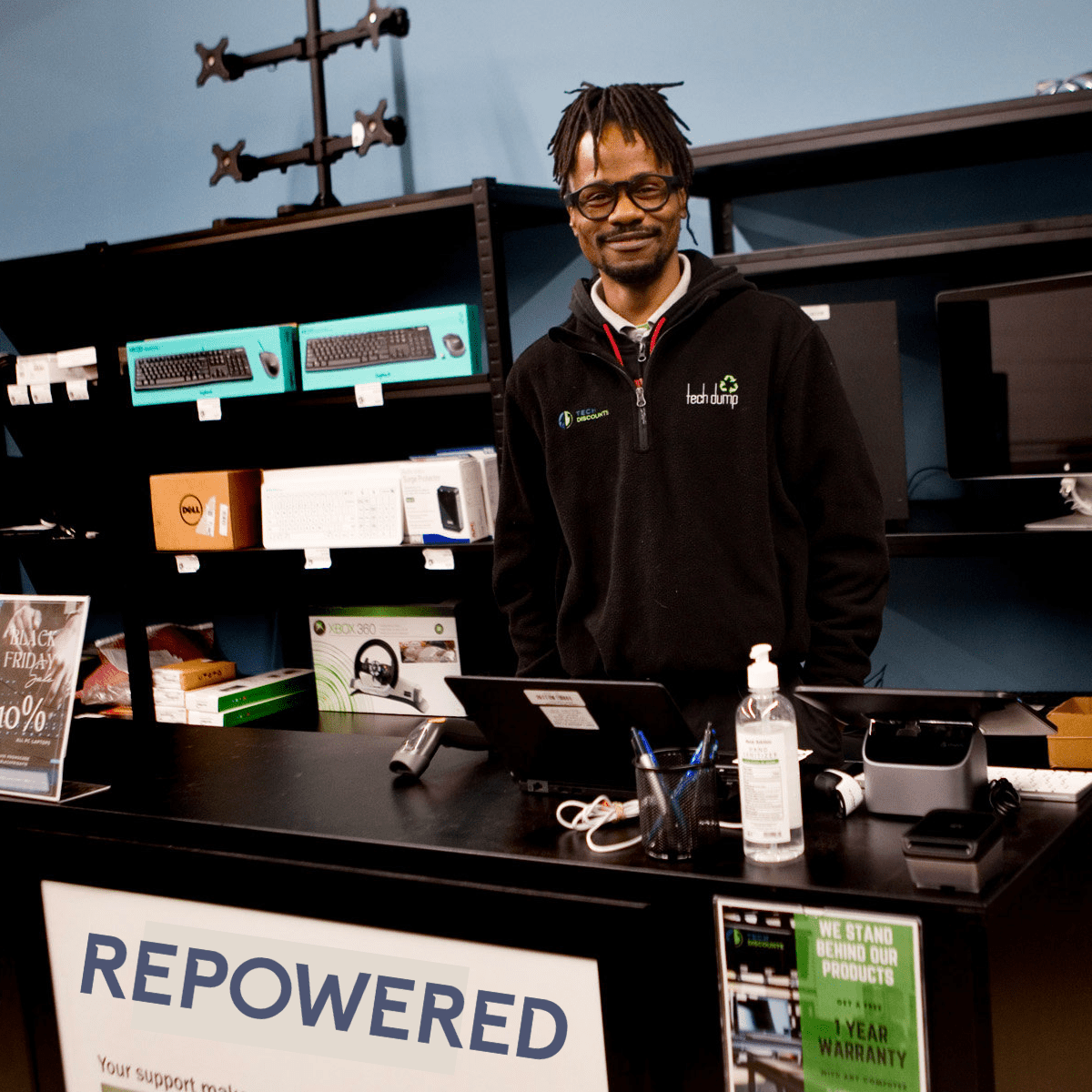 A Black man with braids smiles at the camera from behind a retail counter