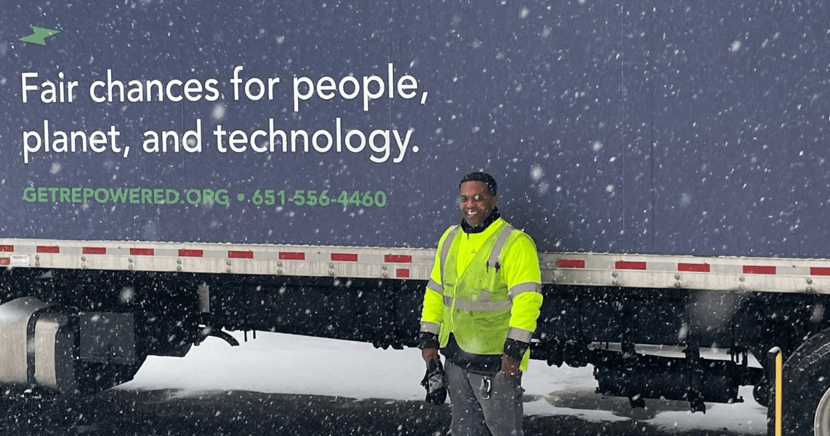 A man standing in front of a truck in the snow smiling at the camera in a reflective vest