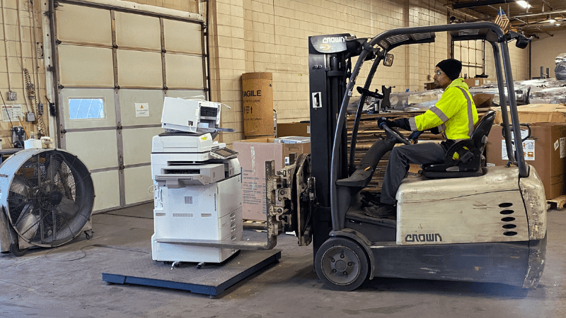 A man in a reflective vest drive a forklift to move a large copy machine
